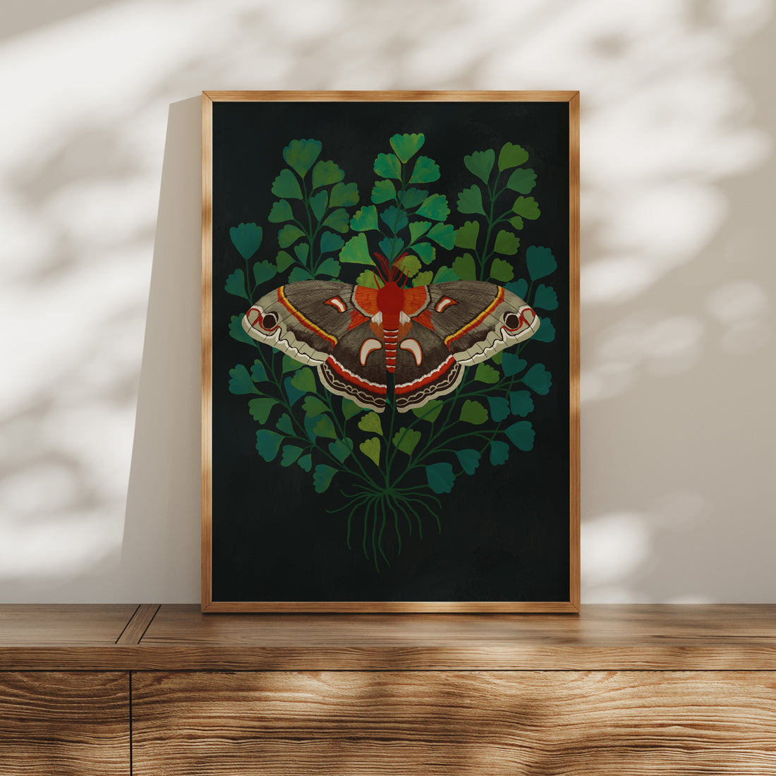 Framed artwork of a butterfly on a black background with green leaves, displayed on a wooden surface.